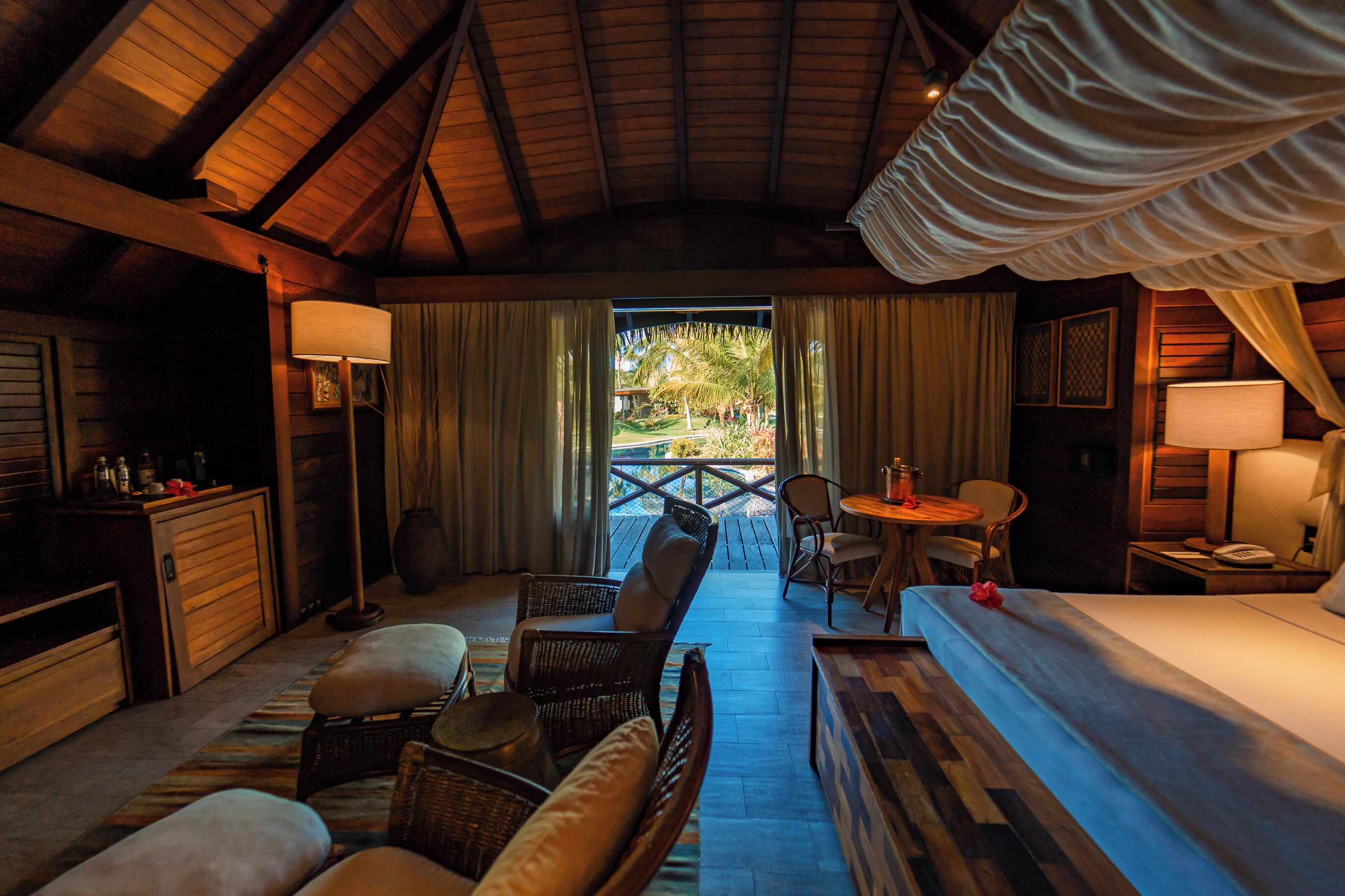 Quarto aconchegante com cama, escrivaninha, cadeira e mesinha. Há uma janela com cortinas, luminária e vista para o exterior.