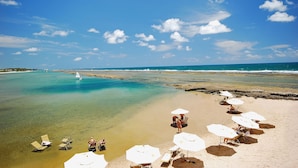 A beach with clear water, a sailboat, and several sun umbrellas and lounge chairs. - NANNAI Muro Alto (Ipojuca)