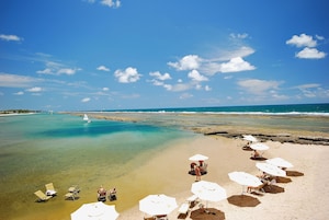A beach with clear water, a sailboat, and several sun umbrellas and lounge chairs.
