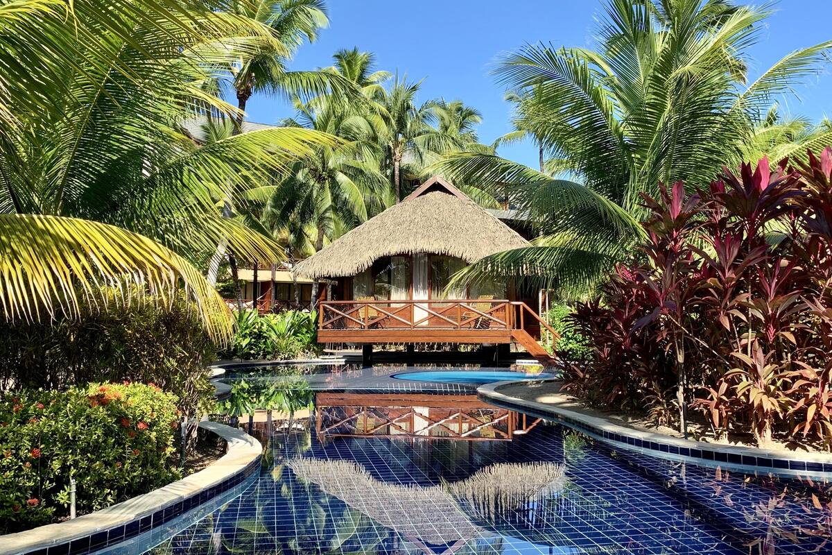 A resort with a pool, palm trees, and a thatched-roof building.
