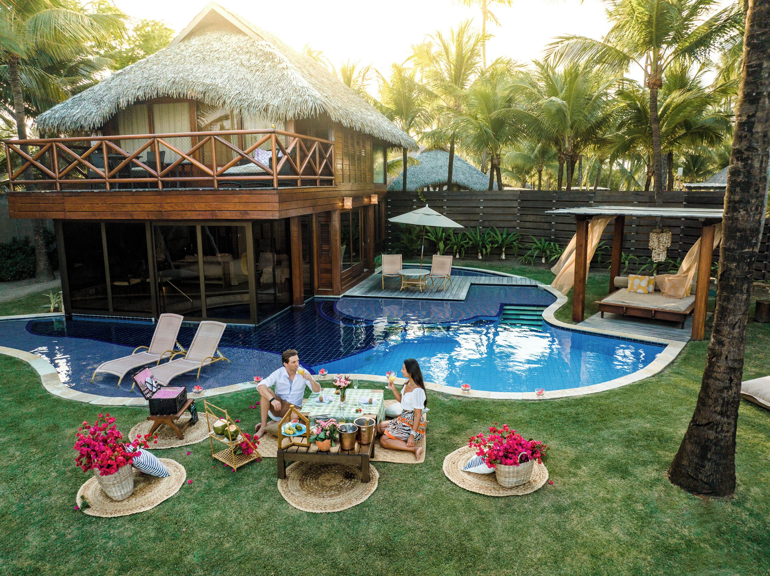 A poolside area with a thatched-roof structure, lounge chairs, and a dining setup with a table and chairs.