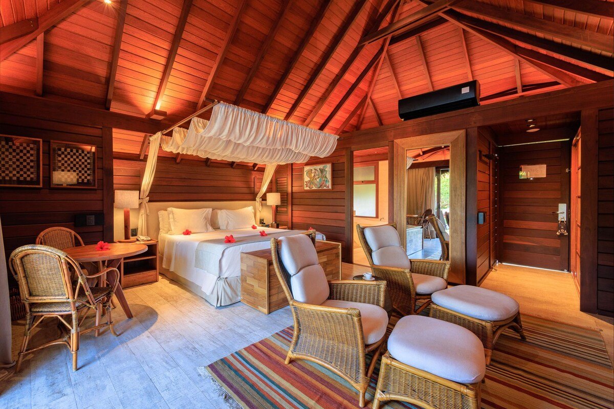 A cozy room with a bed, wicker chairs, a dining table, and a ceiling fan.
