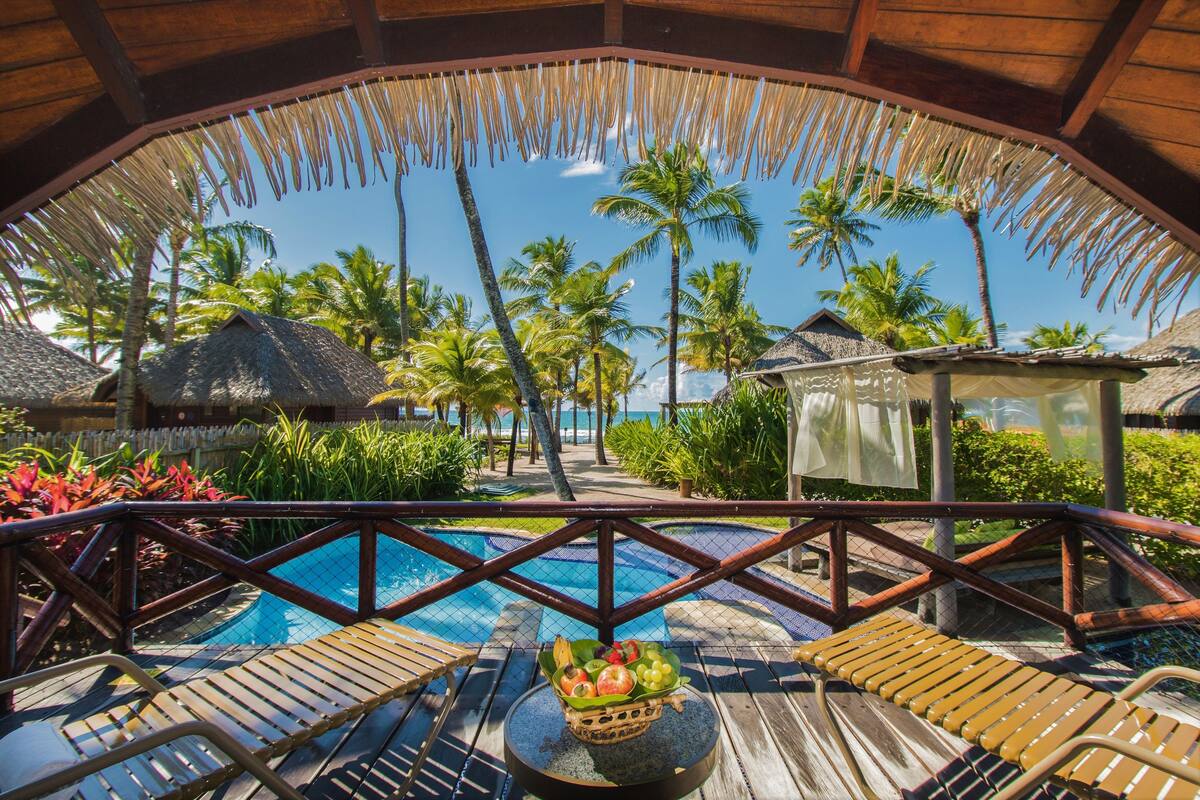 A wooden deck with a pool, surrounded by palm trees and a thatched roof structure.