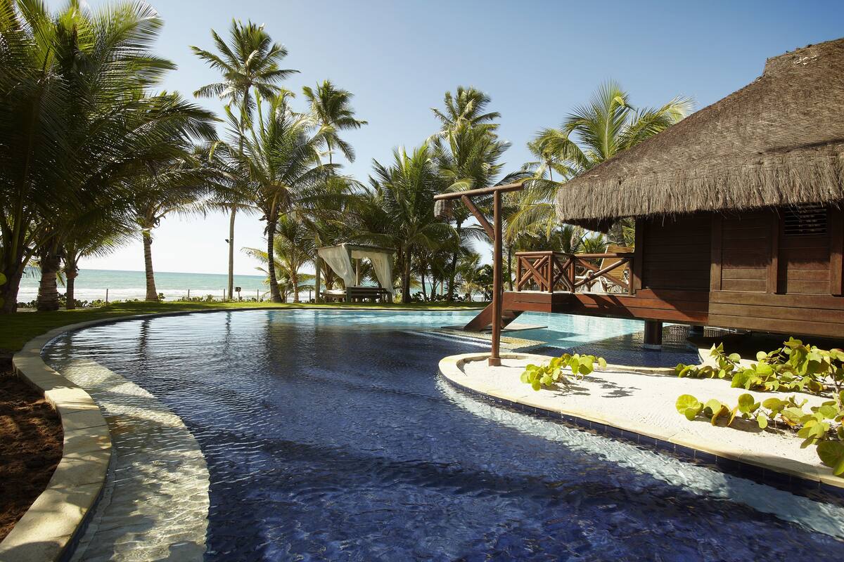 A swimming pool with a thatched-roof structure, palm trees, and a view of the ocean.
