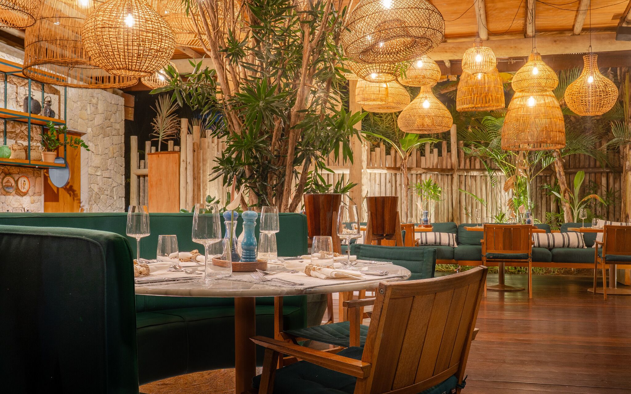Un comedor interior con iluminación colgante de mimbre, asientos tapizados en verde y un fondo de pared de piedra.