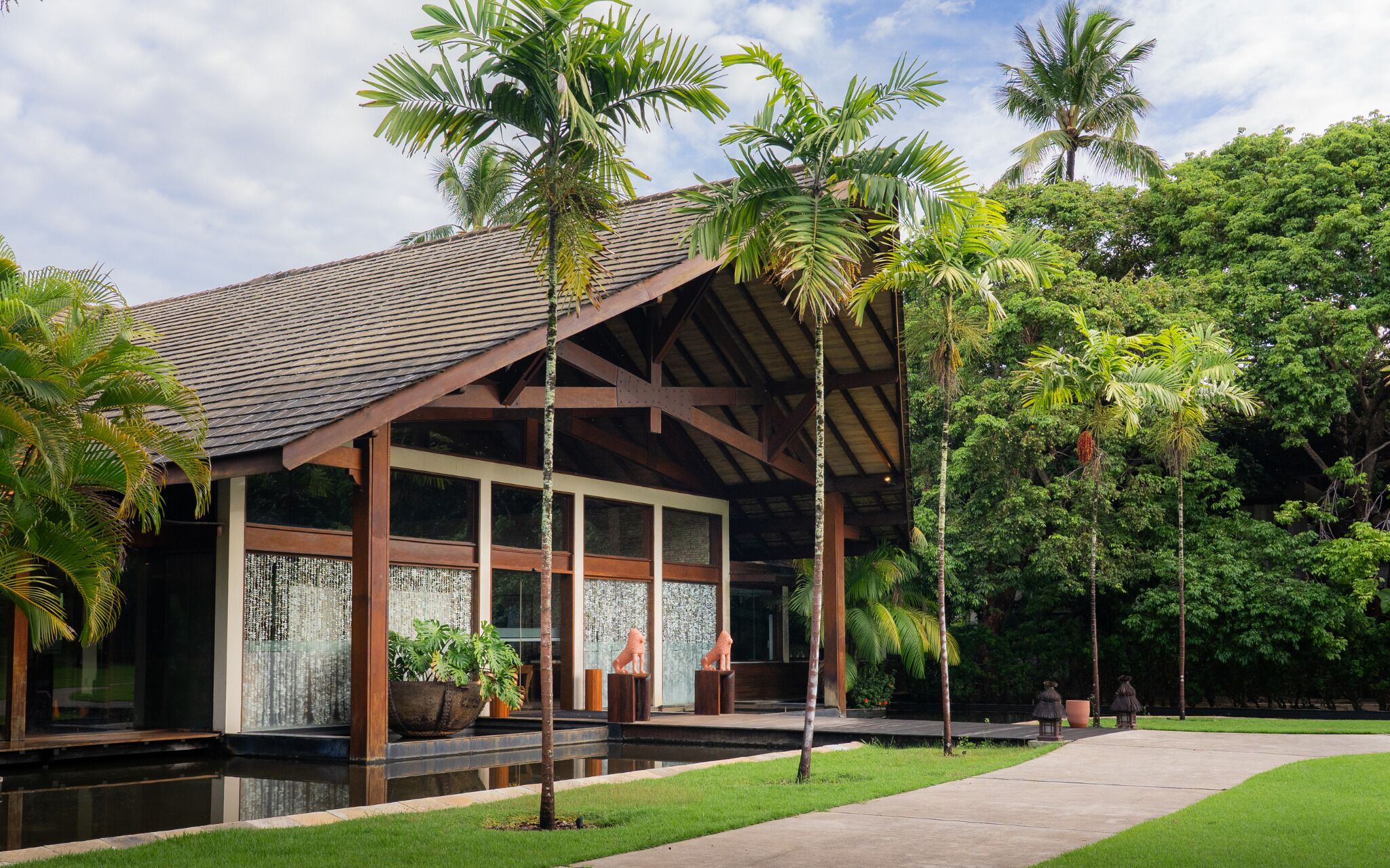 A modern building with a sloped roof, large windows, and a surrounding garden with palm trees.