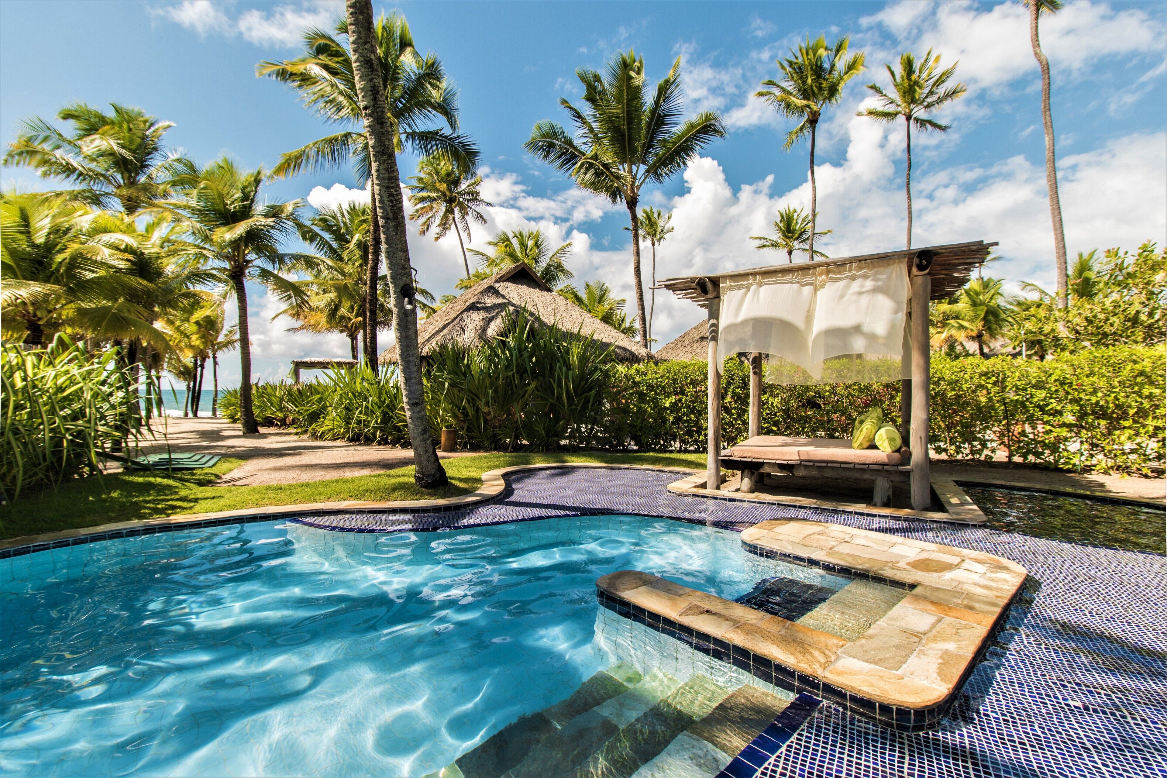 Una piscina con borde de azulejos, rodeada de palmeras y una estructura con techo de paja.
