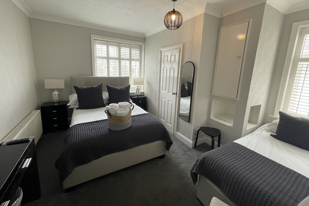 A bedroom with two beds, a bedside table with a lamp, a small table with a lamp, a mirror, and a window with blinds.