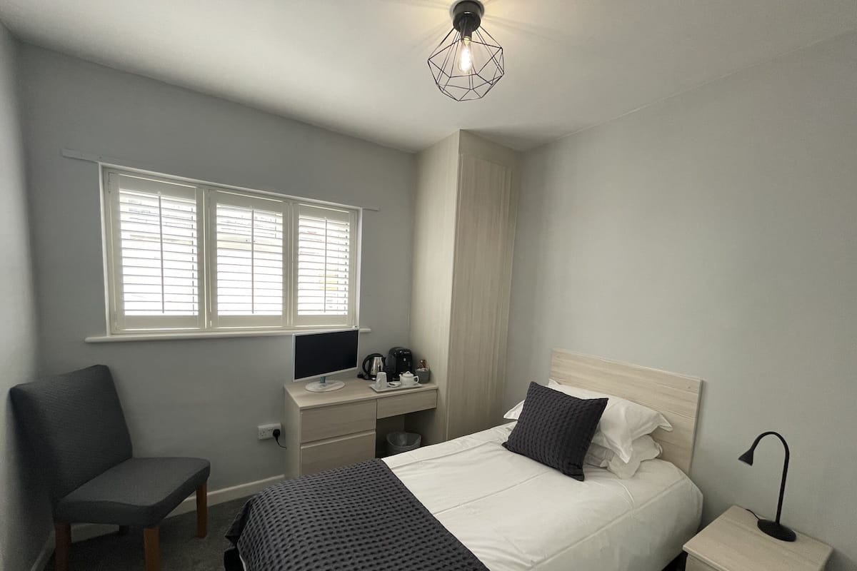 A bedroom with a bed, bedside table, lamp, chair, desk, computer, and window with blinds.