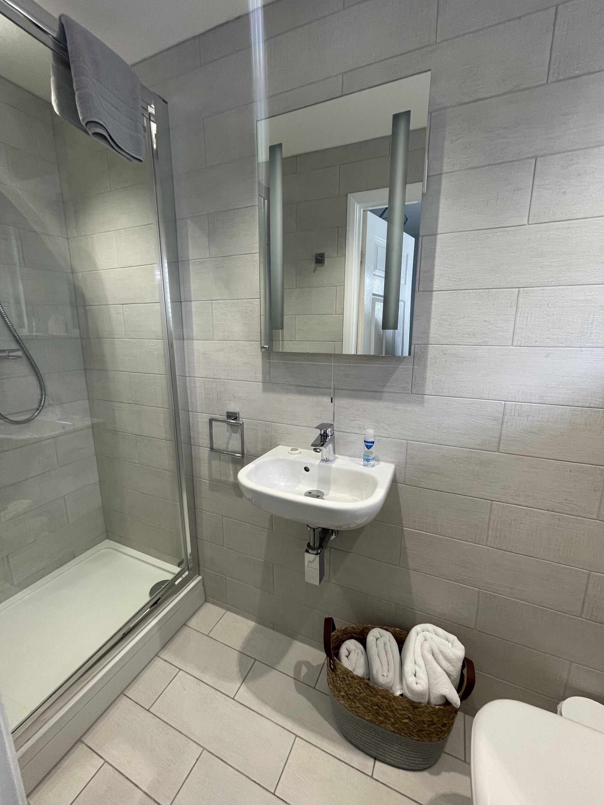 Een badkamer met een glazen douchecabine, een wit wastafeltje met spiegel erboven en een mand handdoeken op de vloer.