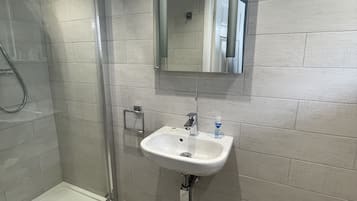 Een badkamer met een glazen douchecabine, een wit wastafeltje met spiegel erboven en een mand handdoeken op de vloer.