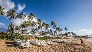 A beach with white lounge chairs and umbrellas, palm trees, and a lifeguard tower. - Catussaba Suítes Resort (Salvador)