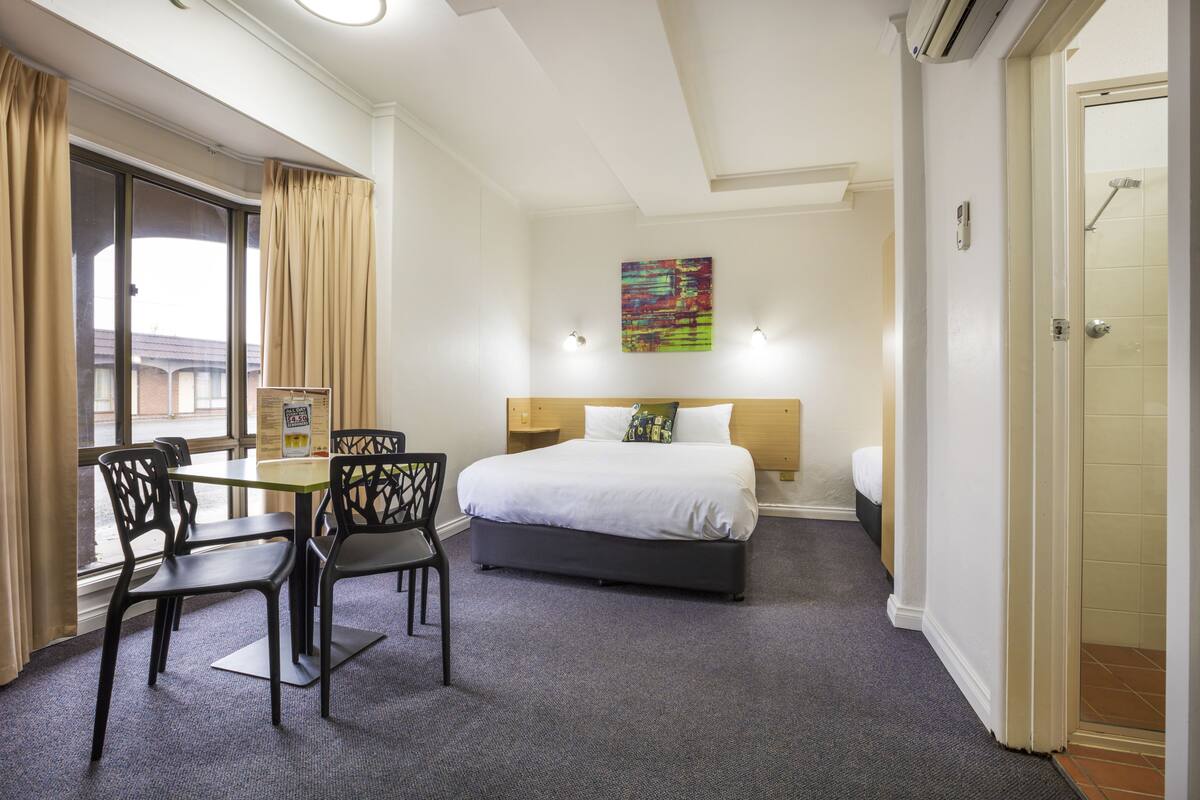 Hotellihuone, jossa on sänky, työpöytä tuoleineen, ikkuna verhoineen, seinällä taulu ja ovi, joka johtaa kylpyhuoneeseen.