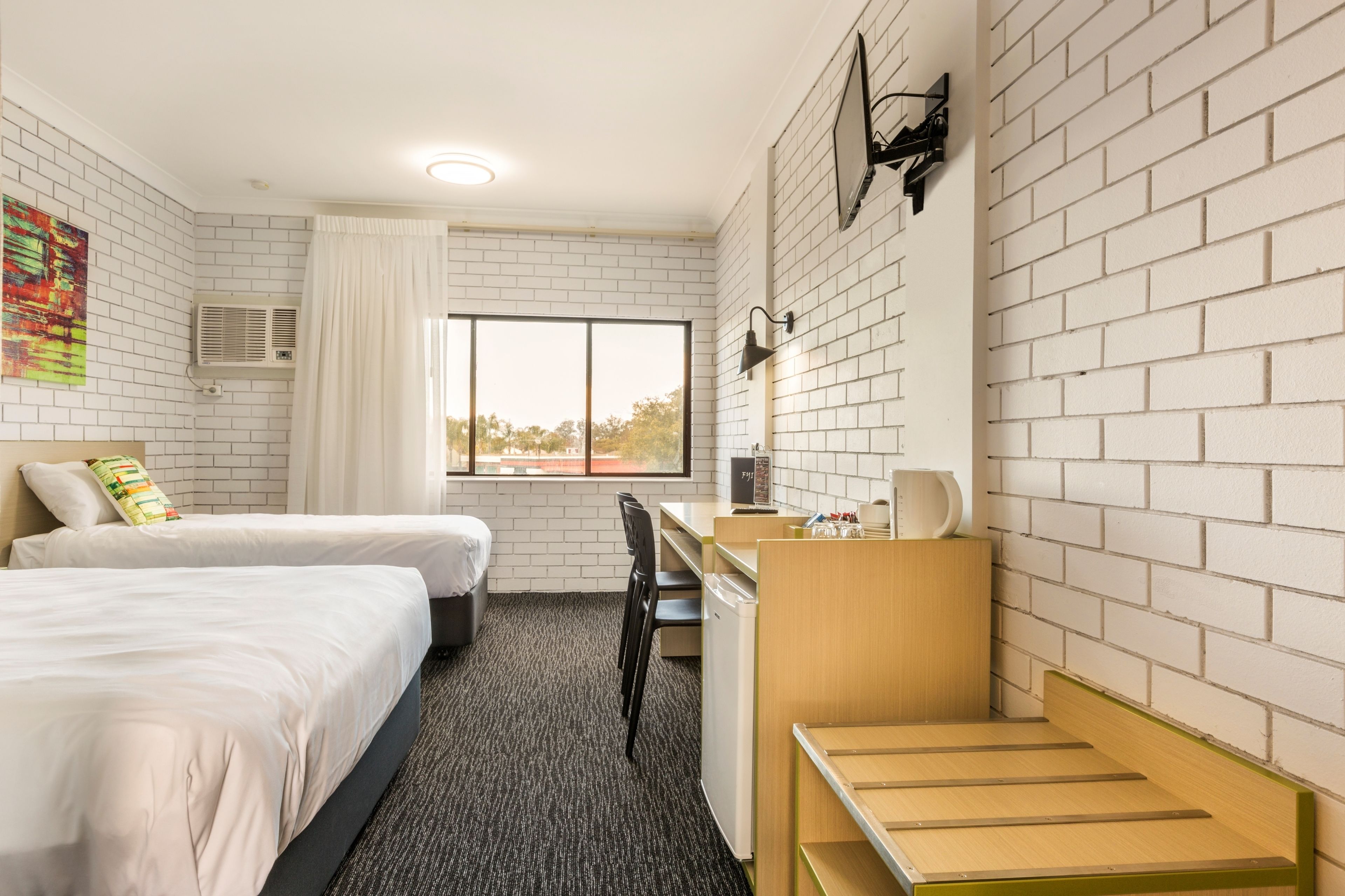 A hotel room with two beds, a desk, a chair, a small refrigerator, and a window with a view.