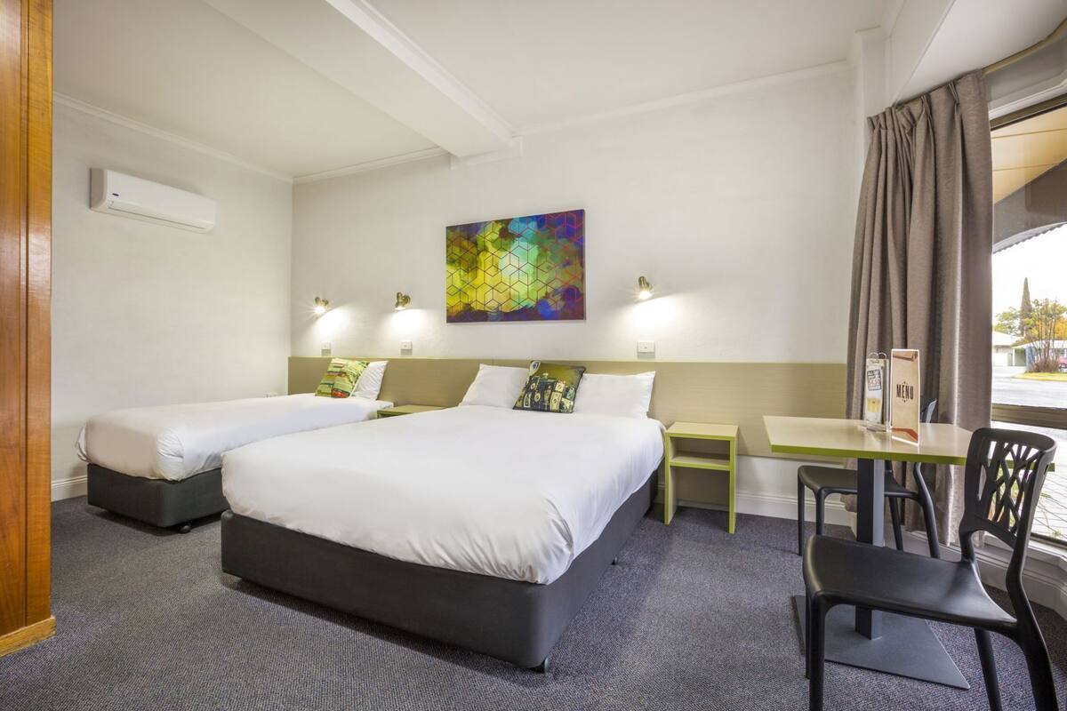 Hotellihuone, jossa on kaksi sänkyä, työpöytä, tuoli, taulu ja ikkuna, jossa on verhot.