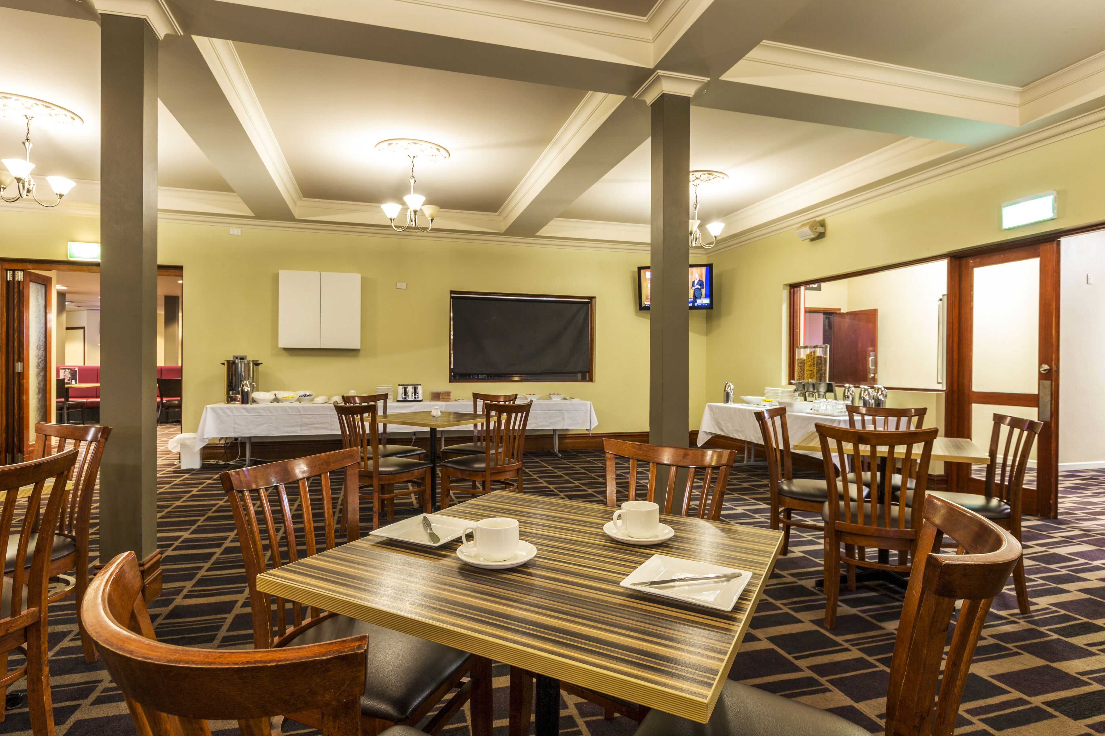 A hotel dining area with multiple tables and chairs, a television screen, and a carpeted floor.