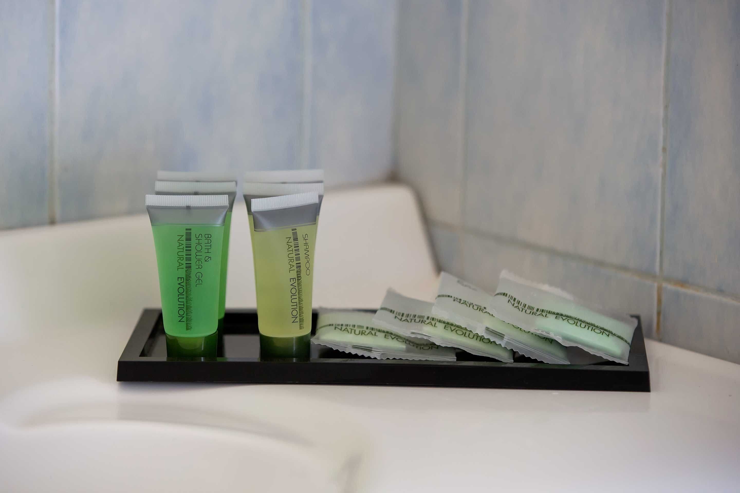 Een badkamer met een zwart dienblad waarop twee groene flessen staan met de tekst 'SHAMPOO & CONDITIONER' en diverse zakjes 'NATURAL EVOLUTION' shampoo.