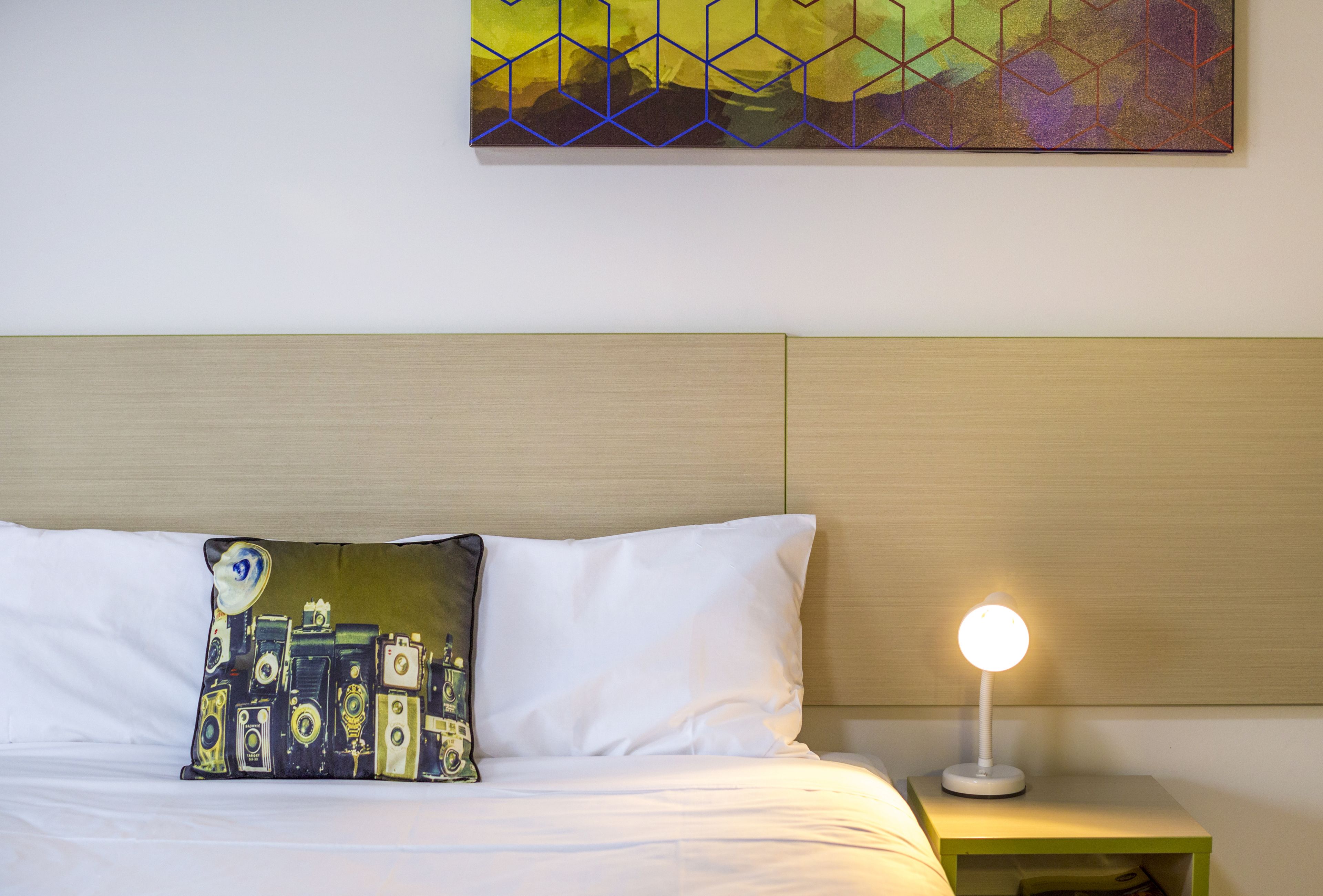 Una cama bien tendida con un almohadón blanco y un cojín decorativo con cámaras antiguas. Se ve una mesita de noche con una lámpara.