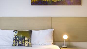 Una cama bien tendida con un almohadón blanco y un cojín decorativo con cámaras antiguas. Se ve una mesita de noche con una lámpara.