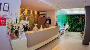 A hotel reception area with a modern design, including a reception desk, framed certificates, and a wall with green foliage. - The Hotel (Salvador)