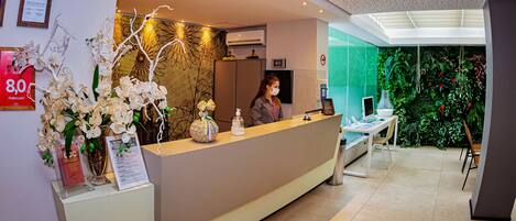 A hotel reception area with a modern design, including a reception desk, framed certificates, and a wall with green foliage.