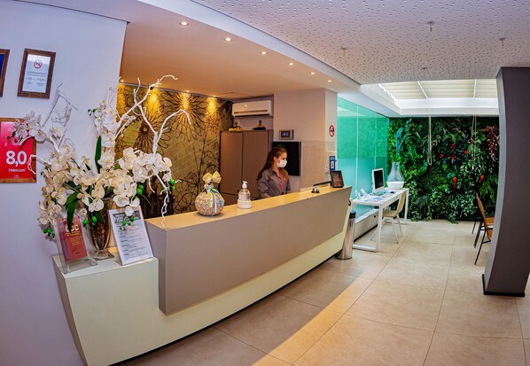 Zona de recepción de un hotel de diseño moderno, con un mostrador de recepción, certificados enmarcados y una pared decorada con follaje verde.