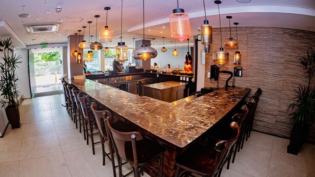 A bar area with a marble countertop, multiple stools, hanging pendant lights, and a wooden accent wall. - The Hotel (Salvador)