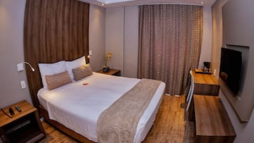 Quarto de hotel moderno com uma cama grande, cabeceira de madeira, televisão de tela plana e uma pequena mesa de cabeceira.
