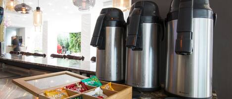 A kitchen area with three stainless steel coffee makers, a wooden box with snacks, and hanging pendant lights.