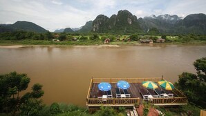 A wooden deck with lounge chairs and umbrellas overlooking a calm river with mountains in the background. - Vansana Vang Vieng (Vang Vieng)