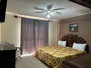 A hotel room with a bed, a desk, a ceiling fan, a window with curtains, and a painting on the wall.
