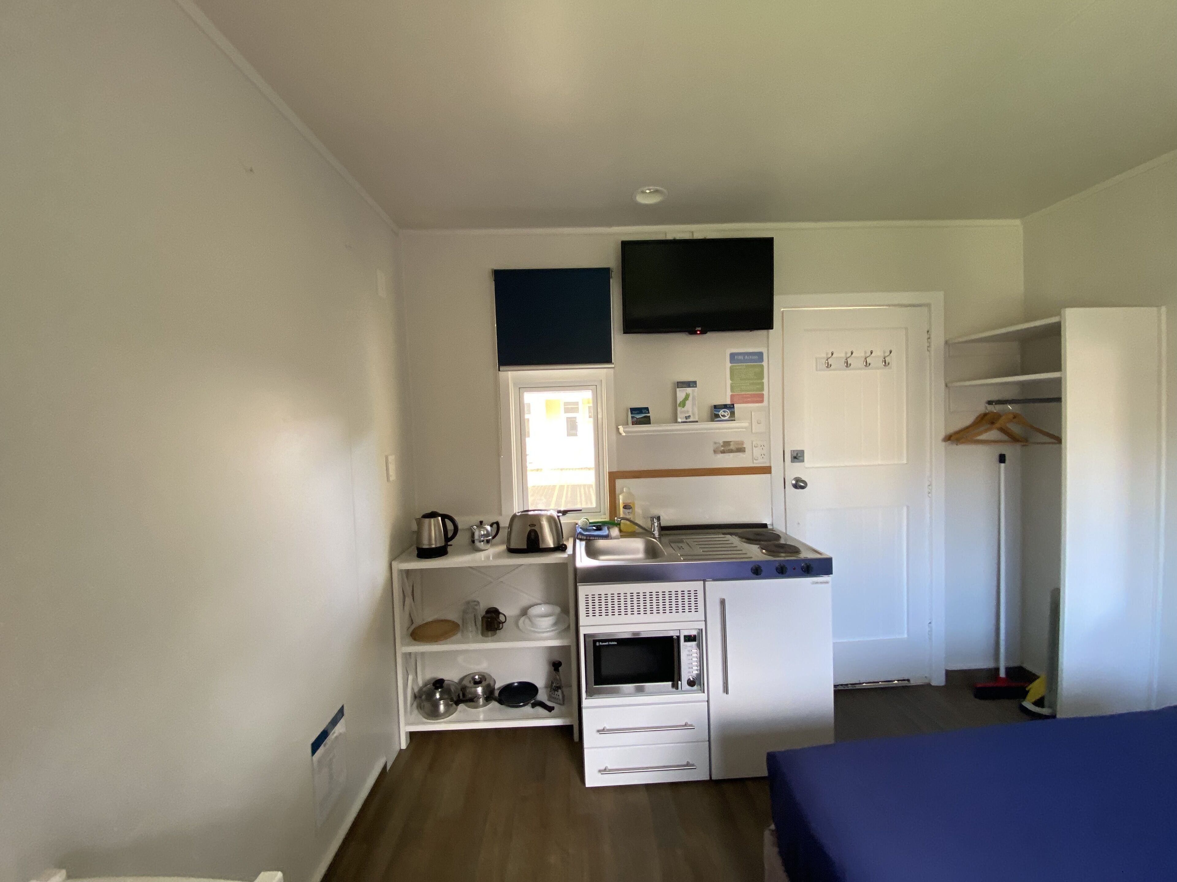 Cocina compacta con microondas, hervidor y cafetera sobre una encimera blanca. Una ventana pequeña proporciona luz natural y un televisor está montado encima de la encimera.