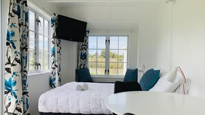 A bedroom with a bed, a TV mounted on the wall, a window with curtains, and a view of the outdoors. - Whanganui River Top 10 Holiday Park (Whanganui)