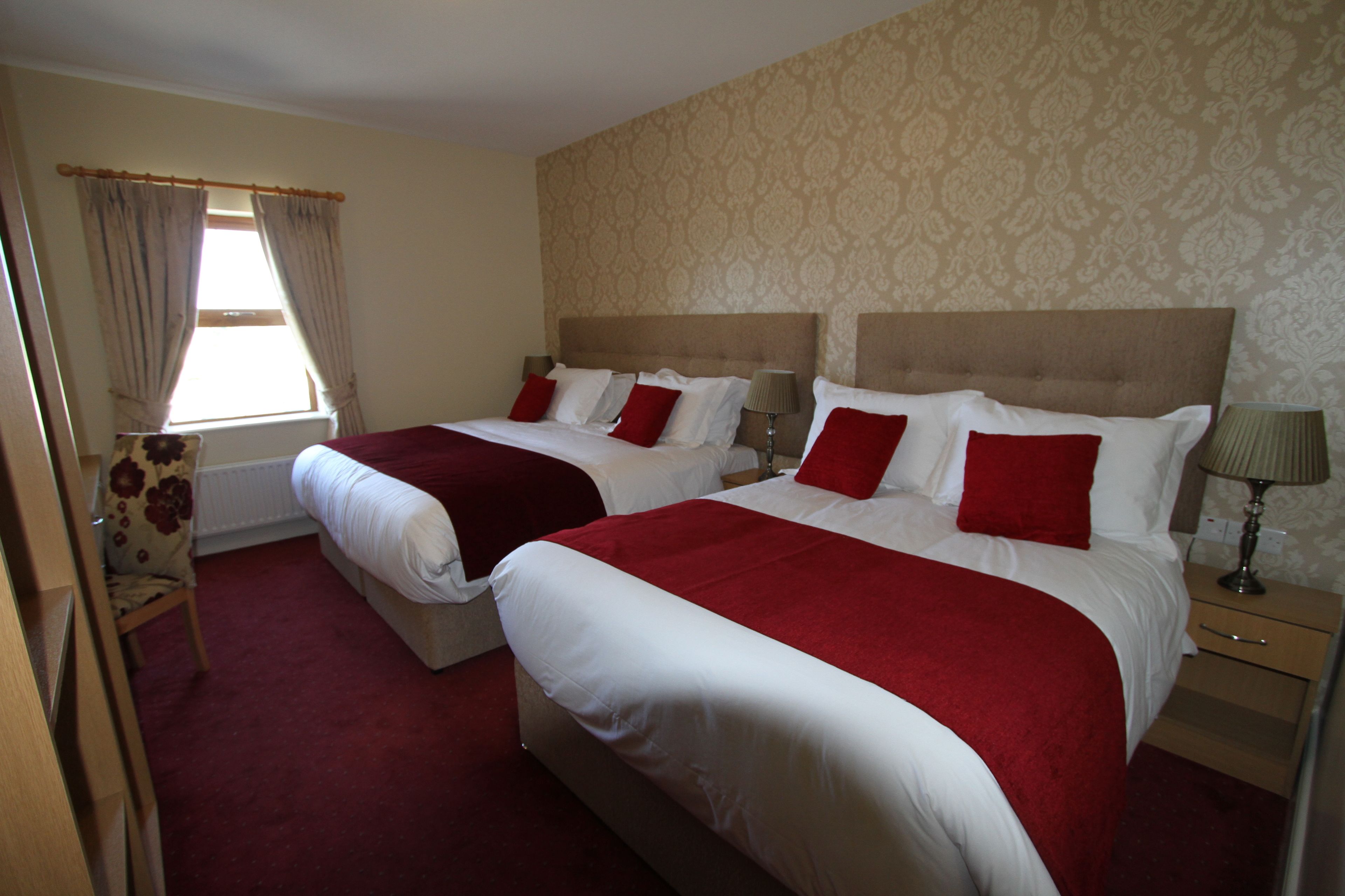 A hotel room with two beds, each with red and white bedding, a wooden nightstand with a lamp, and a window with curtains.