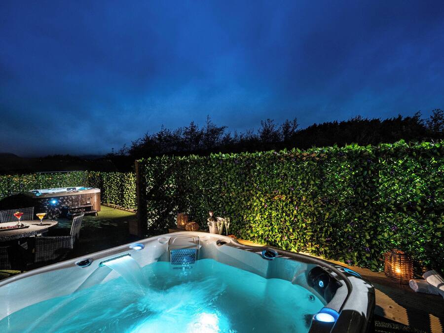A hot tub with a surrounding patio area, including a table and chairs, and a hedge wall.