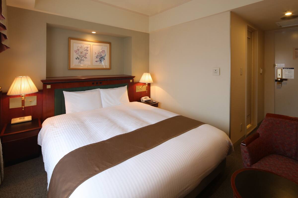 Une chambre d’hôtel avec un grand lit, deux lampes de chevet, une table de chevet, une chaise et une porte donnant sur une autre pièce.