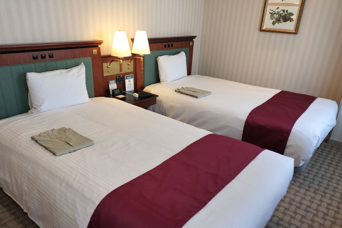 Une chambre d’hôtel avec deux lits, chacun recouvert d’une courtepointe blanche et bordeaux, doté d’une tête de lit verte et agrémenté d’une table de chevet avec une lampe et une horloge.