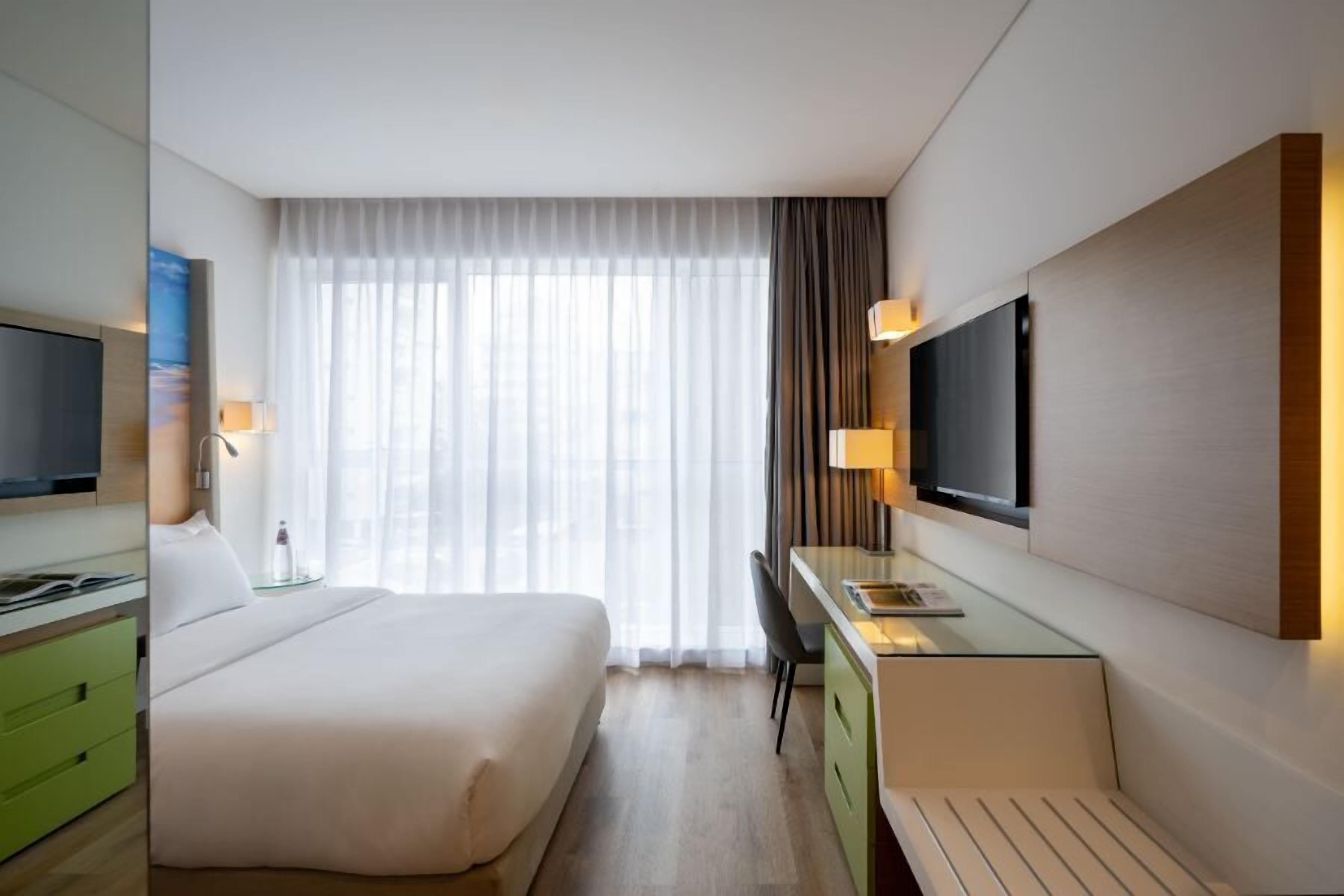 Una habitación de hotel con cama, escritorio, televisión y ventana con cortinas.