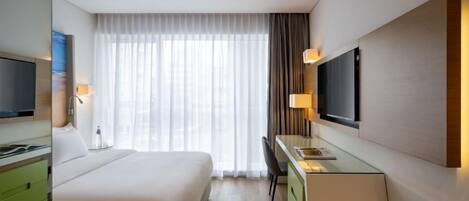 Una habitación de hotel con cama, escritorio, televisión y ventana con cortinas.