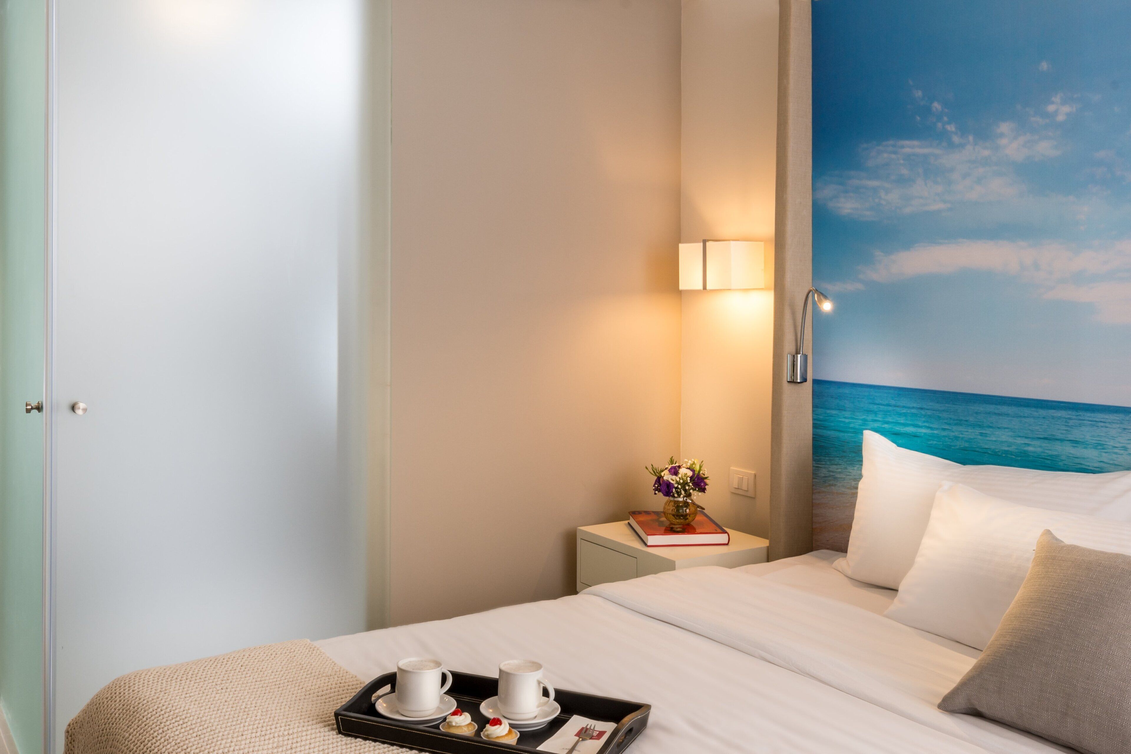 A hotel room with a bed, a nightstand with a lamp, a tray with cups and pastries, and a wall mural of the ocean.