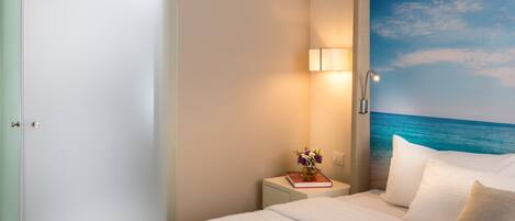 A hotel room with a bed, a nightstand with a lamp, a tray with cups and pastries, and a wall mural of the ocean.