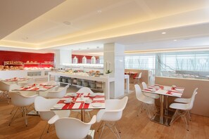 A modern dining area with white tables and chairs, a central counter with food displays, and a view through large windows. - Leonardo Plaza Netanya (Netanya)