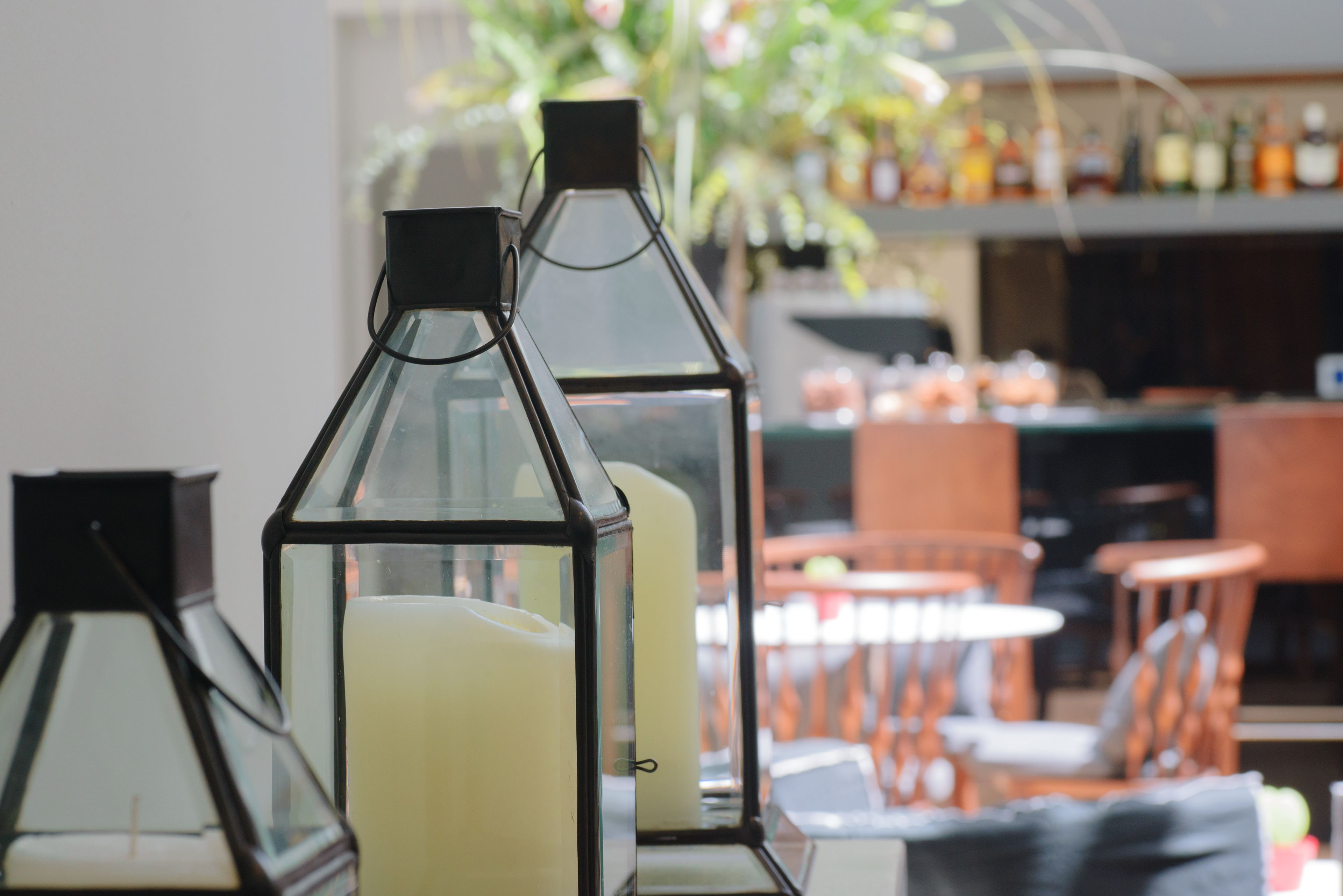A room with a table and chairs, a candle in a glass lantern, and a blurred background with a plant and shelves.