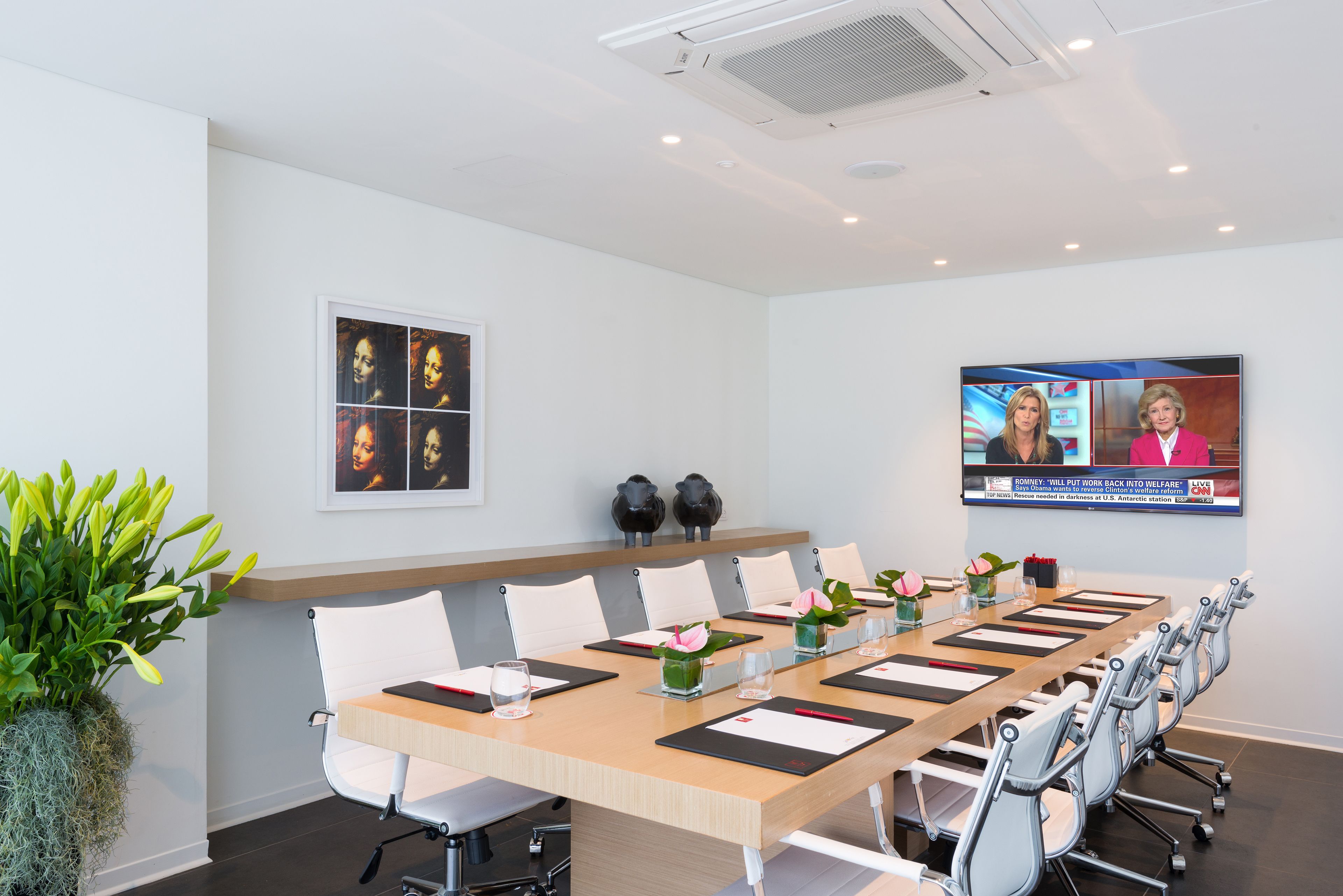 A conference room with a long table, white chairs, and a TV screen displaying a news broadcast.