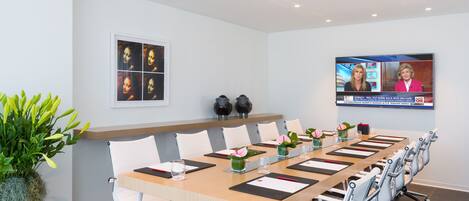 A conference room with a long table, white chairs, and a TV screen displaying a news broadcast.