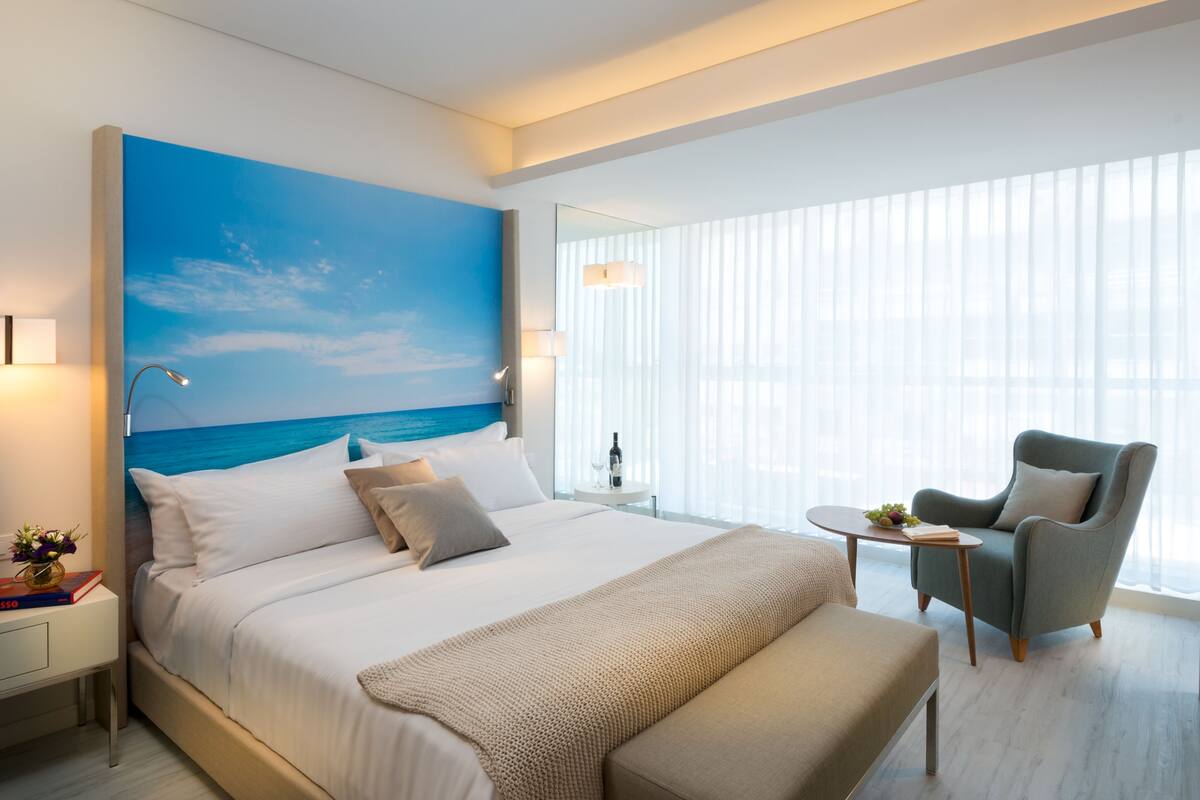 A modern hotel room with a large bed, a view of the sea through the window, a small round table with a bottle and glasses, and a comfortable armchair.