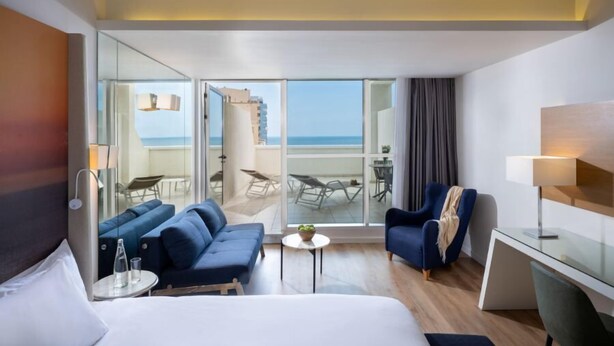 Habitación de hotel con cama, dos sofás, escritorio, silla, mesita y balcón con vista al mar.