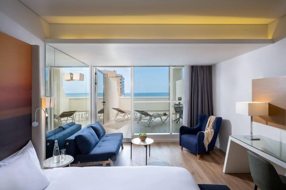 A hotel room with a bed, two sofas, a desk, a chair, a small table, and a balcony with a view of the sea.