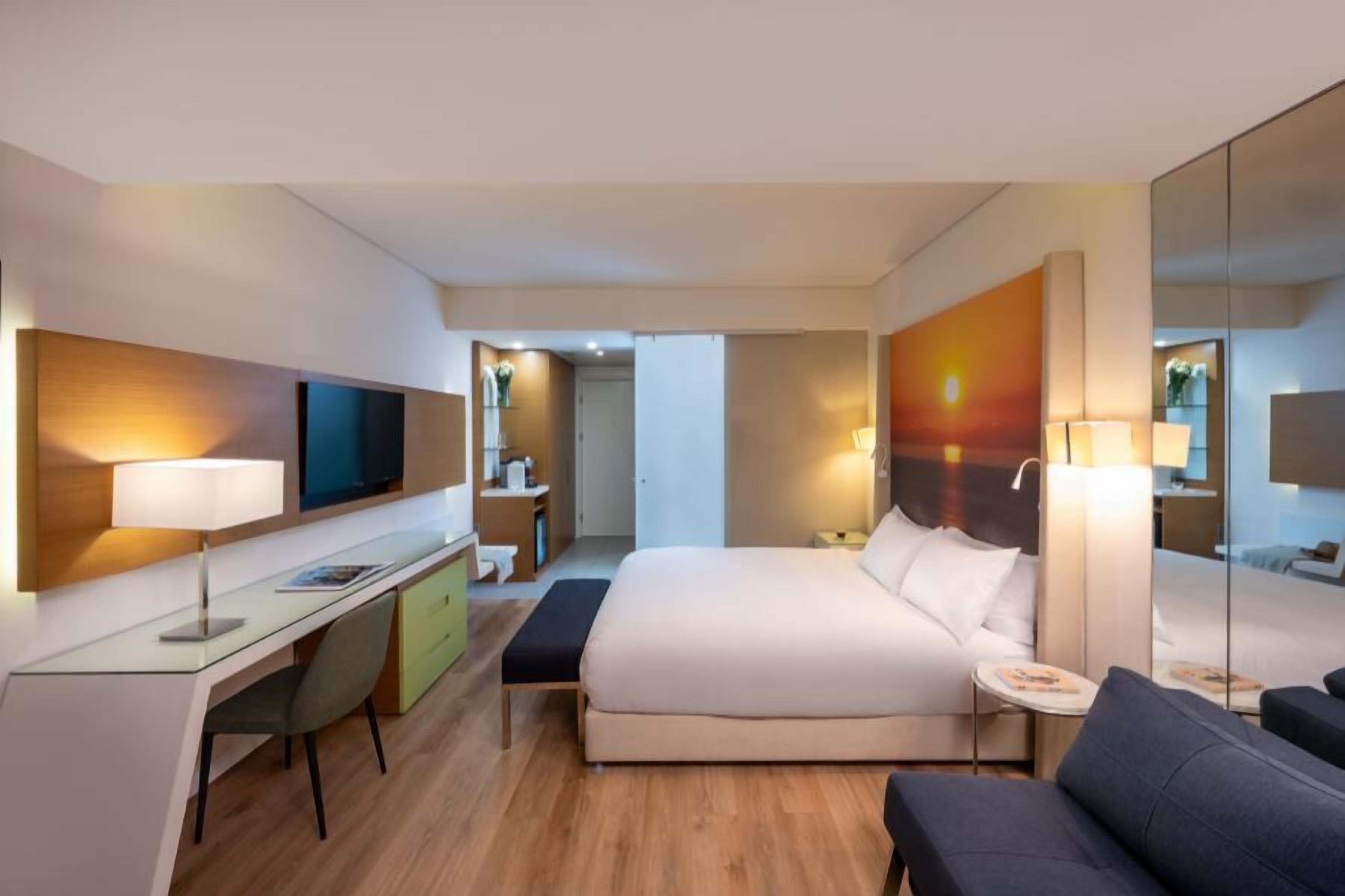 Et moderne hotelværelse med seng, skrivebord, stol, sofa, fjernsyn og spejl.