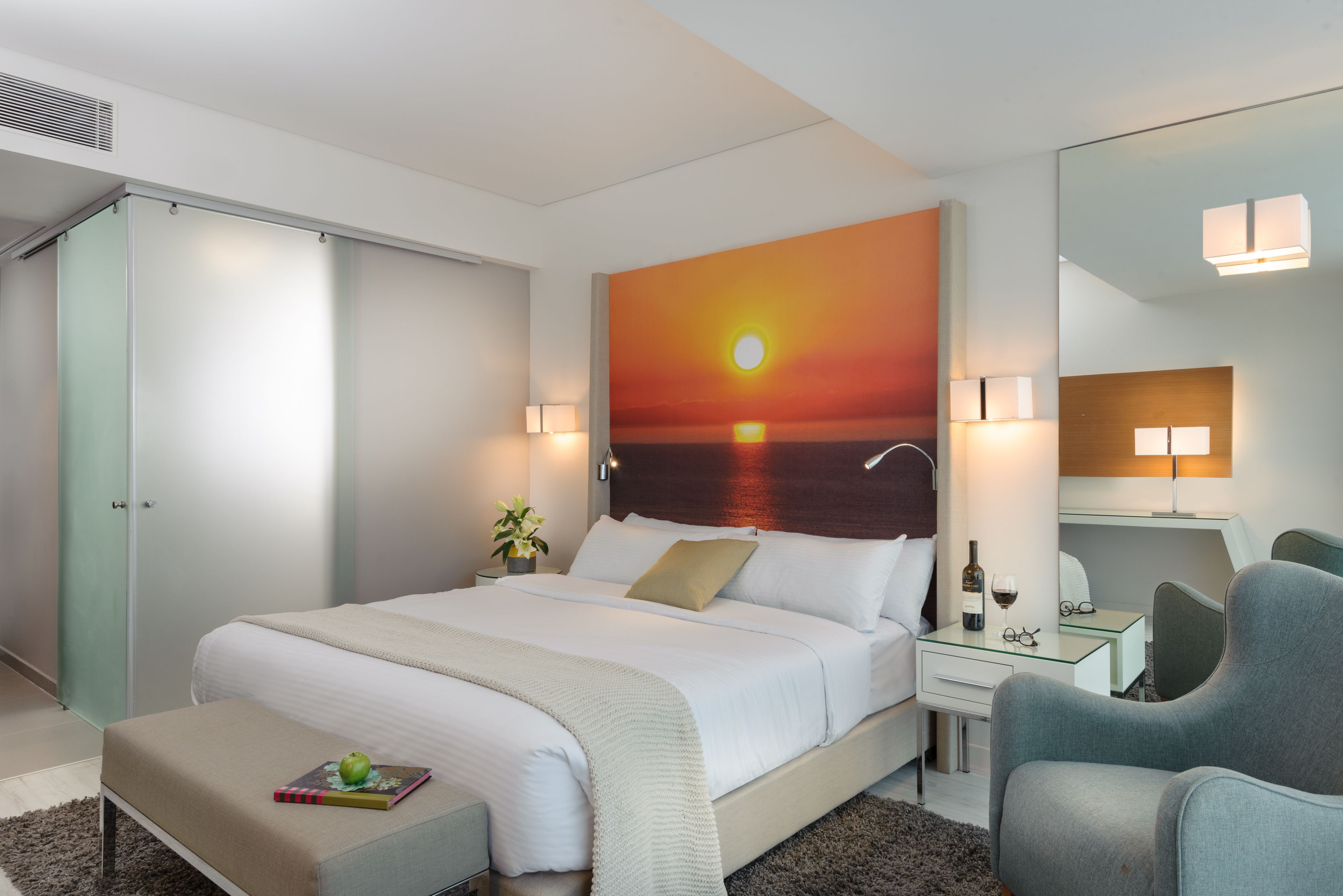 A modern hotel room with a large bed, a desk, a chair, and a painting of a sunset.